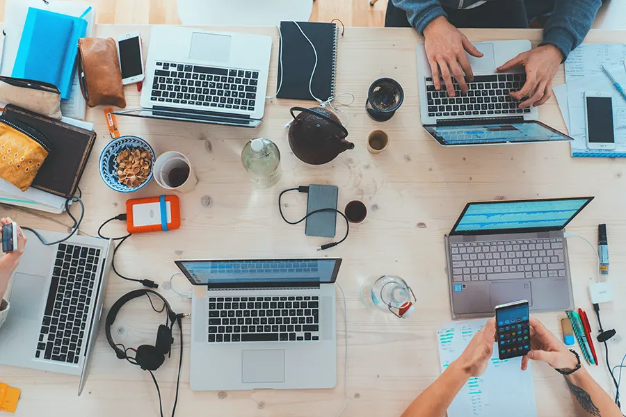 a wooden table is viewed top down where it is covered in laptops, coffee, and cables, appearing to be a table for lots of productivity.